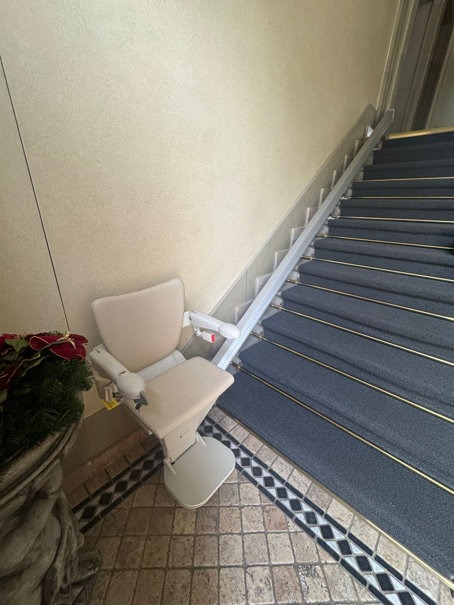 LSM Mobility Un siège monte-escaliers Handicare 1100 beige est replié au bas d'un escalier recouvert de moquette bleue, sur un sol carrelé à motifs. Un mur beige et une plante décorative complètent la scène.