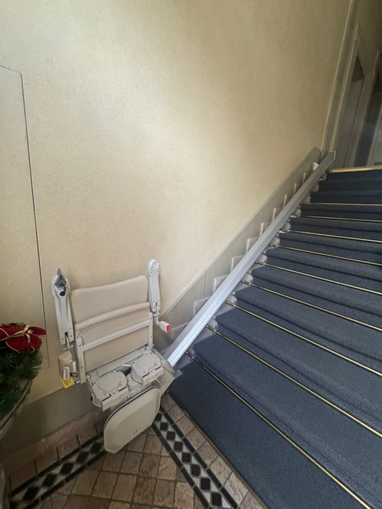 LSM Mobility Siège monte-escaliers Handicare 1100 beige replié au pied d'un escalier intérieur recouvert de moquette bleue avec des bordures dorées. Le rail du monte-escalier longe le mur beige.