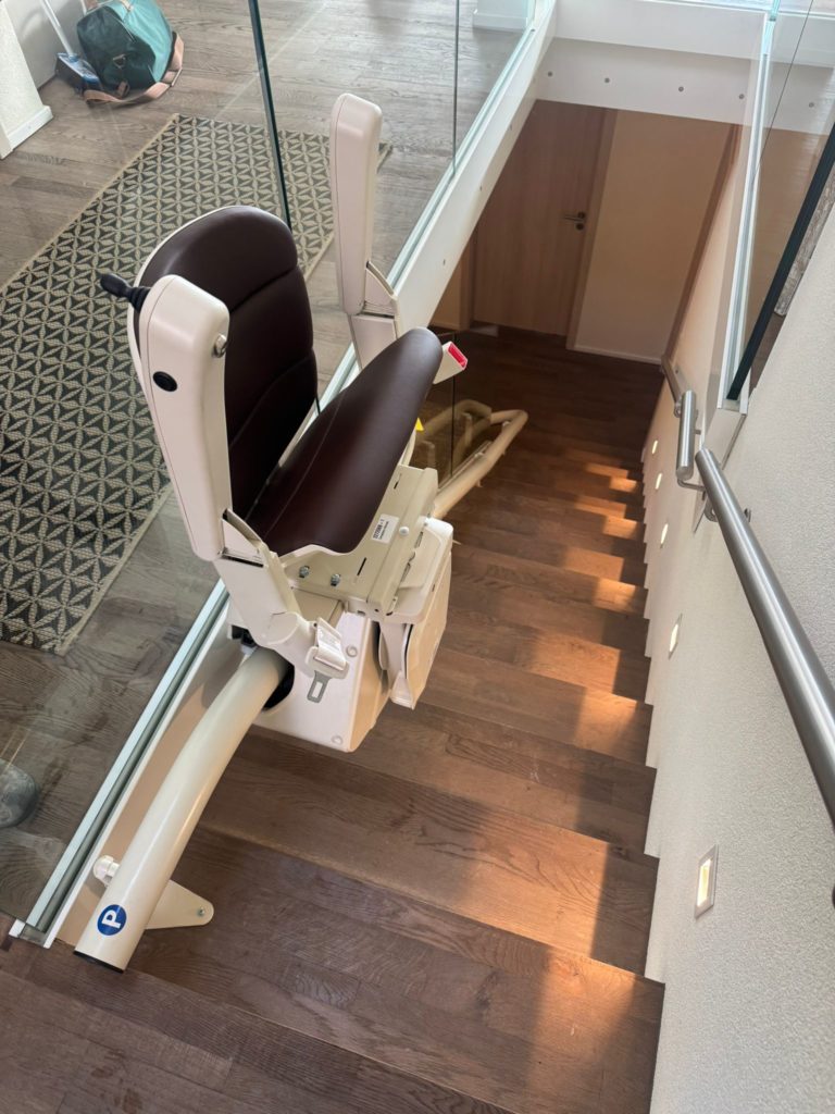 LSM Mobility Un monte-escalier Handicare monorail courbe marron et beige installé sur un escalier en bois sombre avec une rampe en verre. La chaise est repliée, prête à l'emploi.