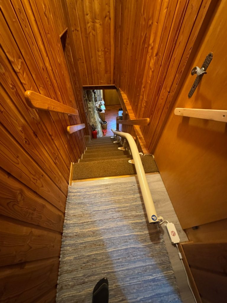 LSM Mobility Vue plongeante sur un escalier en bois avec un siège monte-escalier monorail installé. Le couloir est étroit et entièrement recouvert de bois, menant à une pièce lumineuse au fond.