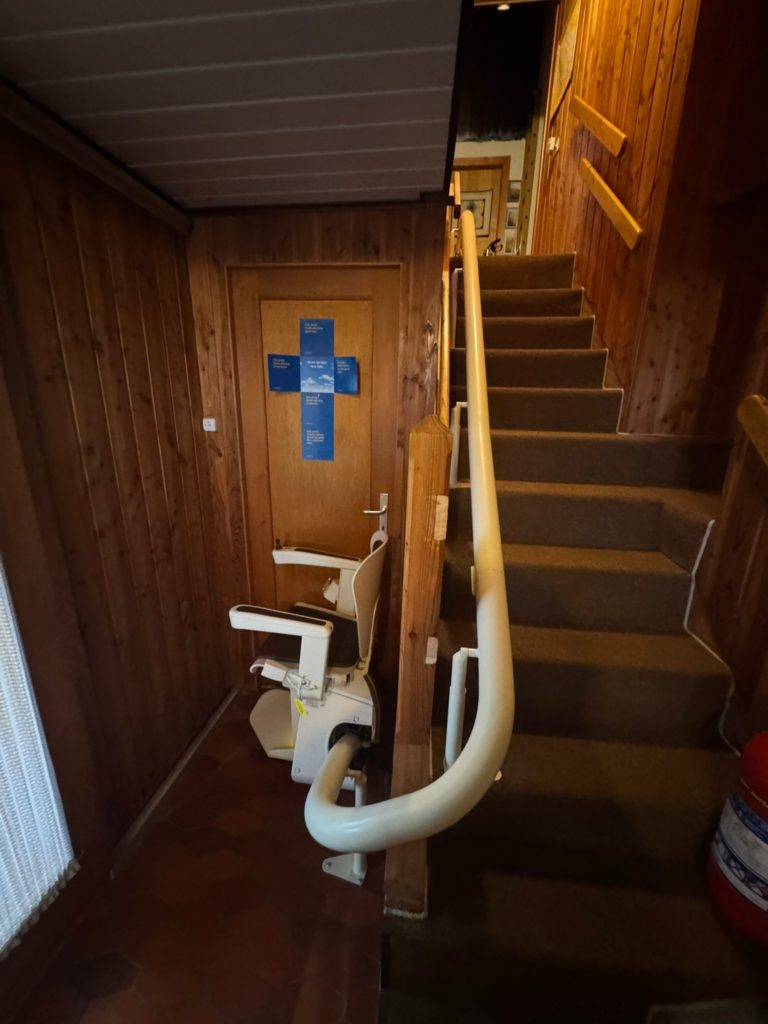 LSM Mobility Intérieur avec escalier en bois équipé d'un siège monte-escalier monorail blanc, facilitant l'accès aux étages. Une porte bleue avec des informations est visible à gauche.