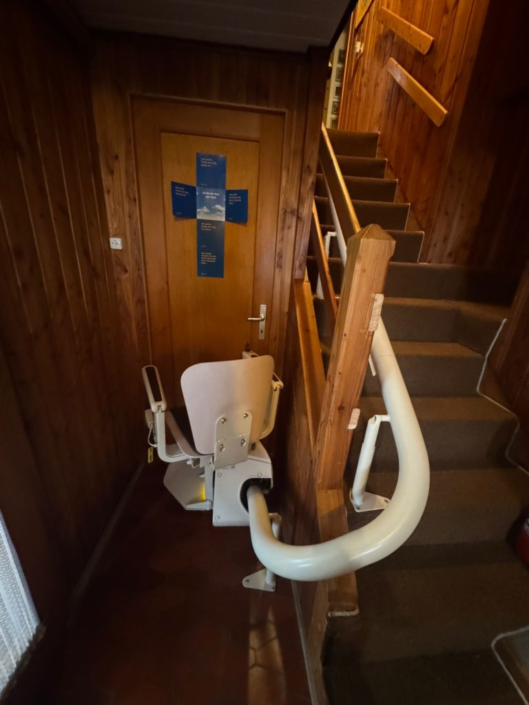 LSM Mobility Siège monte-escalier monorail installé dans un escalier en bois avec moquette marron. Une porte en bois est visible avec des affiches bleues. L'appareil facilite l'accès aux étages.