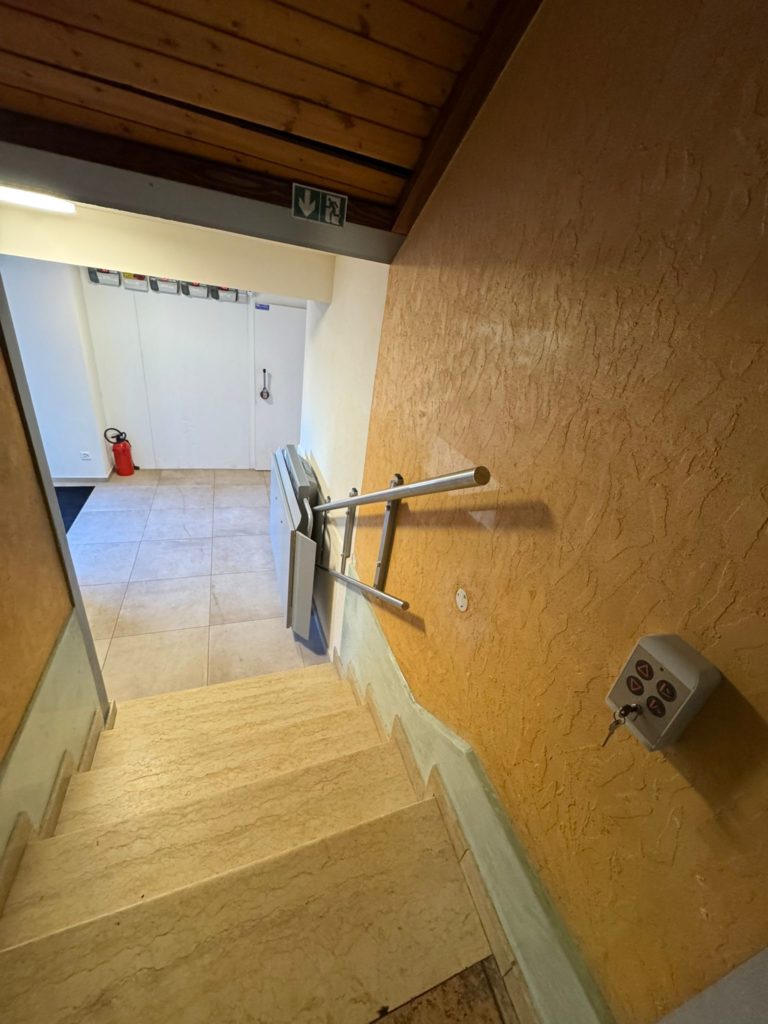 LSM Mobility Vue plongeante sur un escalier avec une plateforme élévatrice PLG7 installée. L'escalier mène à une porte blanche et un couloir éclairé.
