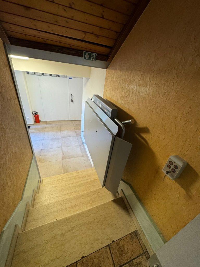 LSM Mobility Plateforme PLG7 installée dans un escalier intérieur. La plateforme élévatrice grise est repliée contre un mur ocre, avec un contrôleur mural à clé visible. Sol carrelé et porte blanche en arrière-plan.