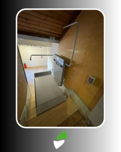 LSM Mobility Plateforme élévatrice PLG7 installée dans un escalier intérieur. La plateforme grise est déployée, prête à l'emploi, avec une rampe de sécurité en métal. Mur ocre et plafond en bois. Accès PMR.