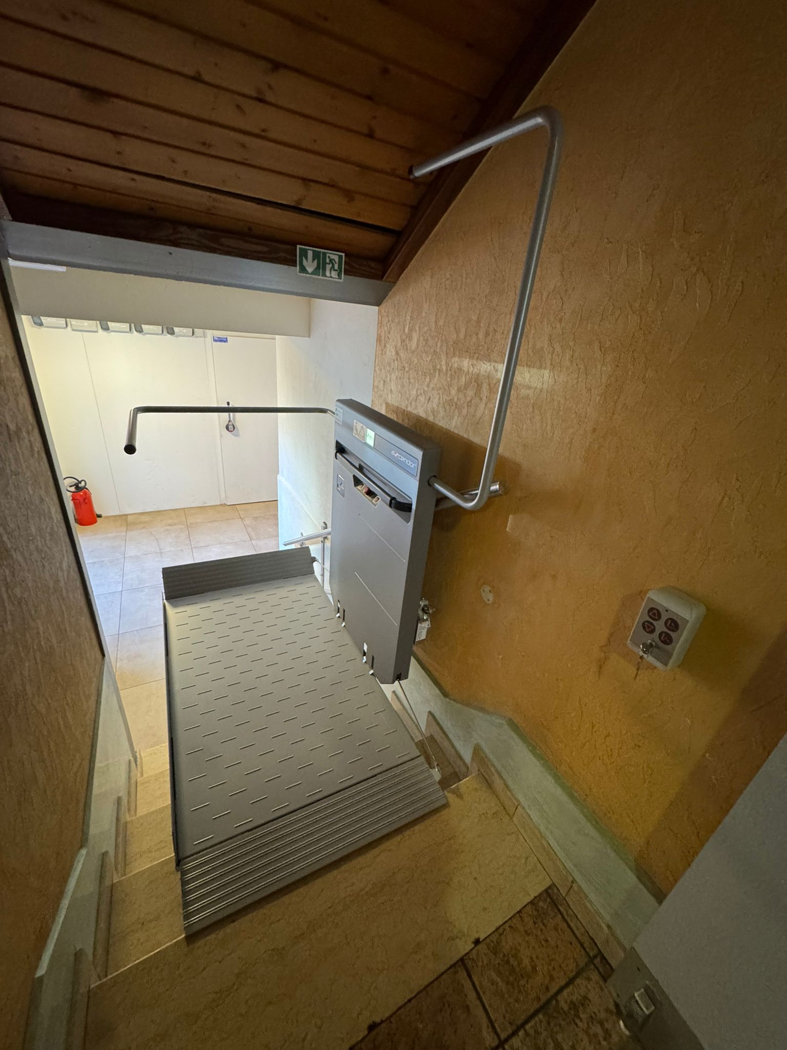 LSM Mobility Plateforme élévatrice PLG7 installée dans un escalier intérieur. La plateforme grise est repliée contre un mur jaune texturé, avec une rampe de sécurité. Un panneau de commande est visible sur le mur.