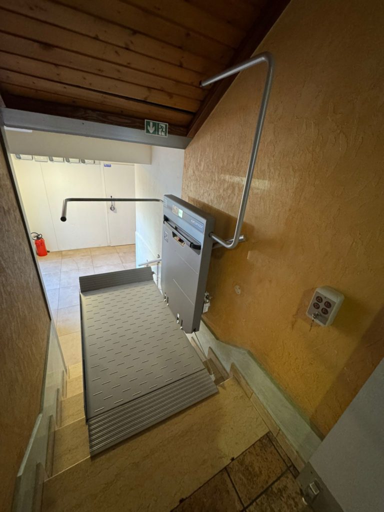 LSM Mobility Plateforme PLG7 monte-escalier gris clair installée dans un escalier intérieur avec murs texturés ocre. Rampe de sécurité argentée. Boutons de commande muraux visibles.