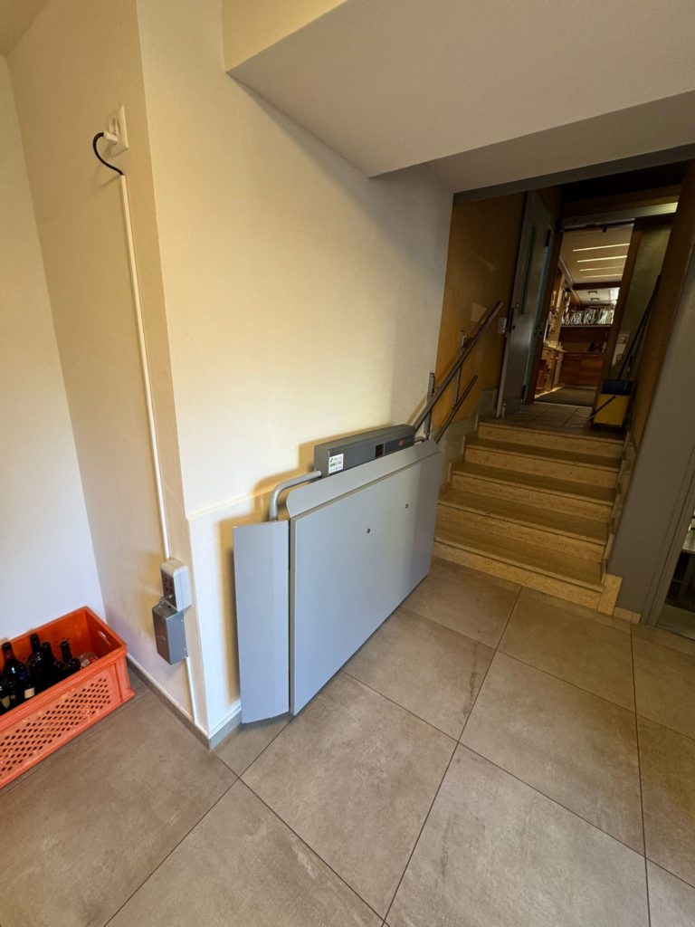 LSM Mobility Plateforme élévatrice PLG7 grise installée dans un couloir menant à un escalier. Une caisse de bouteilles est visible sur le côté. Élévateur pour personnes à mobilité réduite.