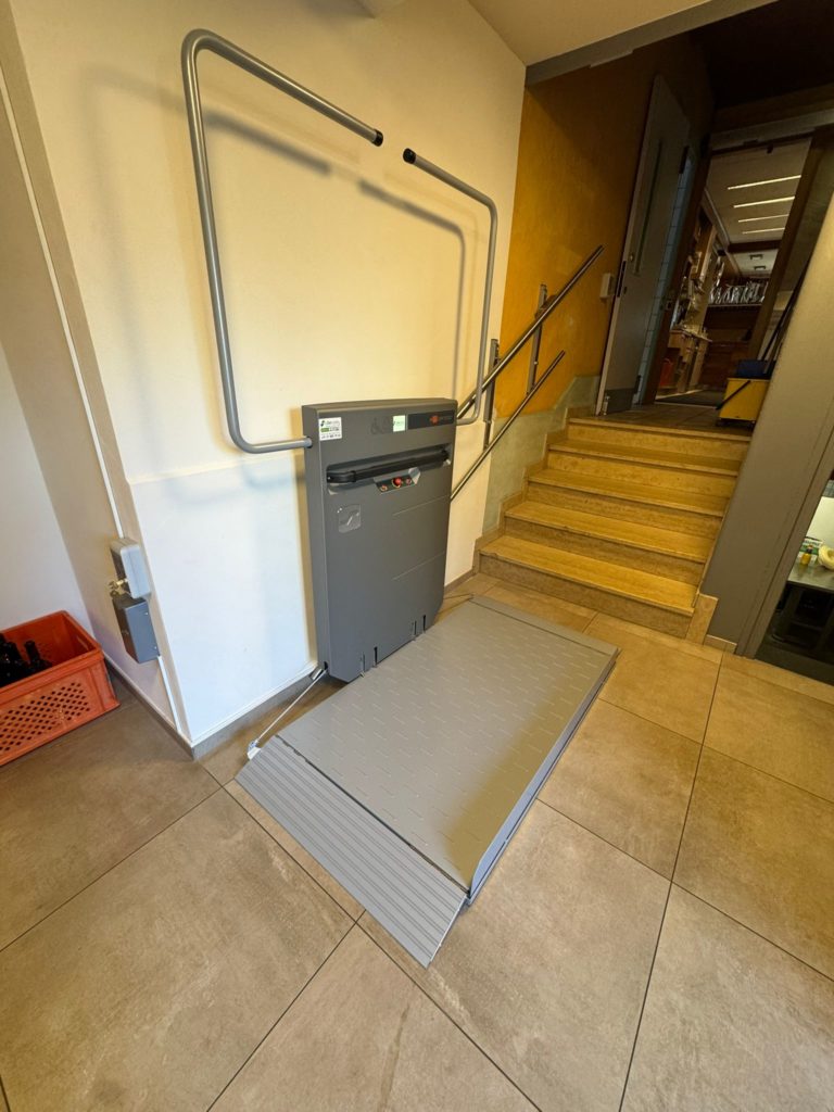 LSM Mobility Plateforme élévatrice PLG7 grise installée près d'un escalier. Dispositif d'accessibilité pour fauteuils roulants, avec rampe et structure de sécurité en métal.