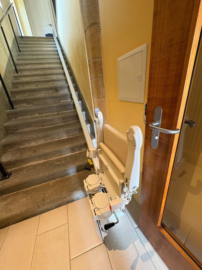 LSM Mobility Siège monte-escalier beige installé sur un escalier en pierre à côté d'une porte en bois.